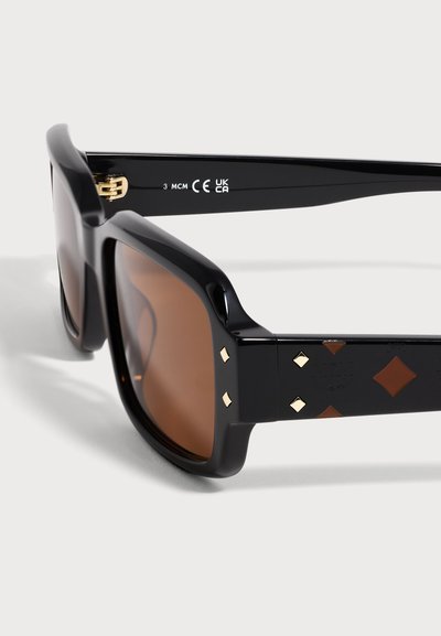 Lunettes de soleil à monture carrée noire avec des verres marron et des détails en diamants dorés et marron sur les branches, présentées en vue rapprochée en angle.