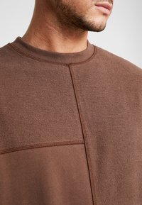 Primo piano della parte inferiore del volto e del collo di un uomo che indossa una felpa marrone in tessuto strutturato e liscio, con dettagli delle cuciture visibili.