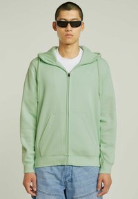 Sweat à capuche zippé vert clair avec capuche, poches avant et poignets côtelés. Porté par-dessus un t-shirt blanc et assorti à un short bleu.