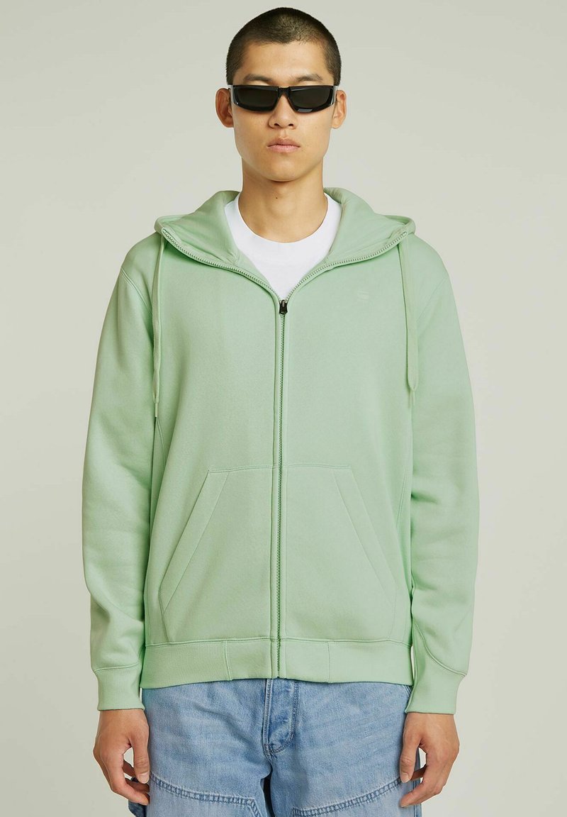 Sweat à capuche zippé vert clair avec capuche, poches avant et poignets côtelés. Porté par-dessus un t-shirt blanc et assorti à un short bleu.