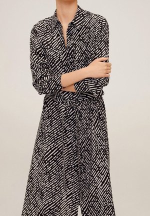 Femme portant une robe-chemise à manches longues avec un col et une ceinture à la taille, en motif noir et blanc, les bras croisés sur la poitrine.