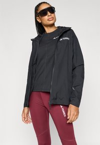 Čierna ľahká bunda so zipsovým uzáverom, nastaviteľnou kapucňou a logovými detailmi. Nosená na priliehavom čiernom tričku a bordových legínach.