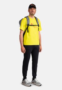 Světle žluté tričko s grafickým potiskem, černé tepláky, šedé běžecké boty a šedý batoh. Černá kšiltovka s logem.