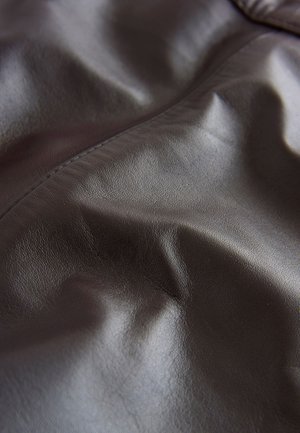 Bruin lederen stof met een glad, gestructureerd oppervlak, met subtiele vouwen en een gelijkmatige glans. De stiksels zijn minimaal en onopvallend.