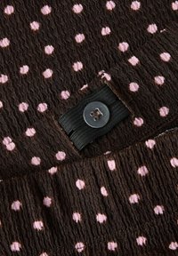 Tissu marron à pois roses, avec une surface texturée. Bande élastique noire avec un bouton métallique en détail sur le bord.