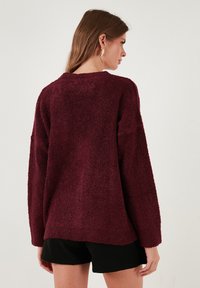 Burgundi színű, bolyhos pulóver texturált anyagból; laza szabású, hosszú ujjú és kerek nyakkivágással rendelkezik. Fekete sorttal viselve.