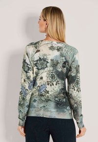 Vrouw met steil blond haar draagt een langarmige top met een bloemen- en abstract patroon in gedempte groene en blauwe tinten, met de rug naar de camera gekeerd.