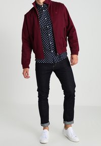Man in een bordeauxrode jacket over een marineblauw polkadot shirt, donkere opgerolde jeans en witte sneakers, staand tegen een effen achtergrond.