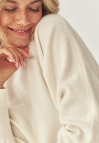 Cremefarvet strikket sweater med en blød tekstur, der har en afslappet pasform og ribkanter, præsenteret mod en lys baggrund.
