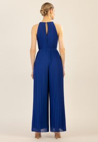 Niebieski kombinezon z plisowaniem, wysokim dekoltem, detalem w kształcie dziurki na plecach, szerokimi nogawkami i wcięciem w talii. Tkanina ma gładką teksturę.