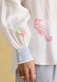 Weißes, leichtes Hemd mit puffigen Ärmeln. Verfügt über rote und grüne Erdbeeren sowie rosa Seepferdchen-Stickereien. Die Manschette hat ein blaues Rüschendetail.