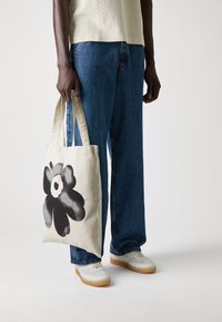 Canvas tote bag featuring a black floral design on a beige background, with sturdy straps and a rectangular shape, held by a person.