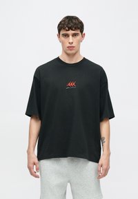 Μαύρο βαμβακερό μπλουζάκι με κόκκινο και λευκό κεντητό σχέδιο στο στήθος. Χαλαρή εφαρμογή με κοντά μανίκια, συνδυασμένο με γκρι σορτς.