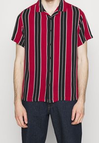 Mężczyzna ubrany w koszulę na krótki rękaw z pionowymi czarnymi, czerwonymi i białymi paskami, w połączeniu z ciemnoniebieskimi dżinsami.