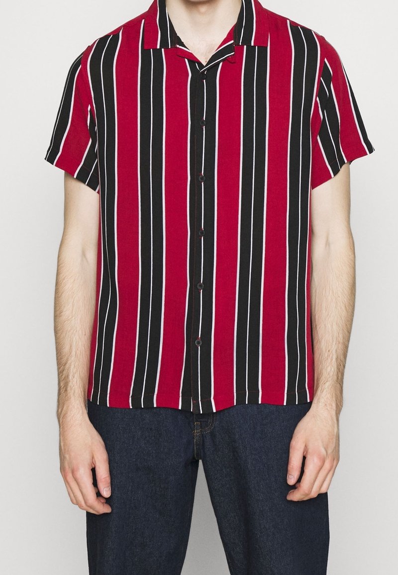 Uomo che indossa una camicia a maniche corte con bottoni, a righe verticali nere, rosse e bianche, abbinata a jeans blu scuro.