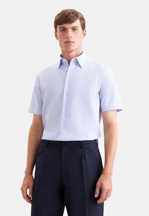 Mężczyzna w jasnoniebieskiej, kraciastej koszuli z krótkim rękawem, włożonej do ciemnogranatowych spodni, stoi na jednolitym tle.