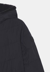 Giubbotto imbottito blu navy con cappuccio, caratterizzato da sezioni trapuntate orizzontali, una morbida fodera in pelliccia sul cappuccio e un logo rotondo sulla manica.