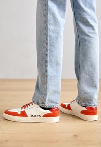 Hvite og oransje sneakers med en buet design, som har en teksturert såle og trykt tekst "HML TN" på siden. Kombinert med lyseblå jeans.