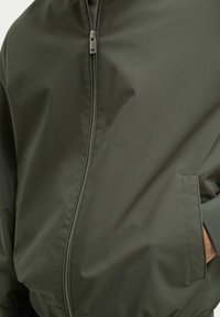 Chaqueta bomber verde oliva hecha de un tejido suave y ligero. Cuenta con un cierre frontal, bolsillos laterales y acentos de puños acanalados.