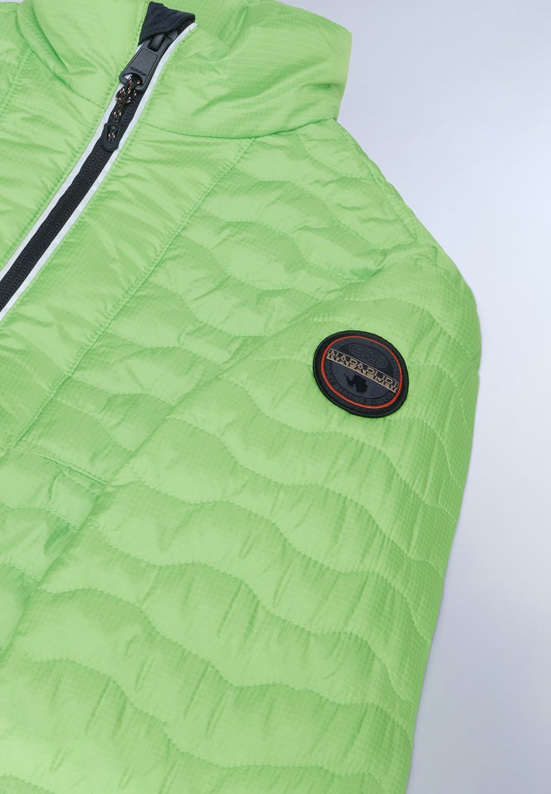 Chaqueta acolchada verde claro con capucha, que presenta un zipper negro, detalles en blanco y un parche negro circular con un contorno rojo en el hombro.