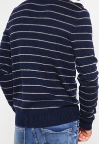 Námořnický pruhovaný svetr vyrobený z měkké látky, s vodorovnými béžovými pruhy, s ribbovaným lemem a dlouhými rukávy, zobrazený zezadu.