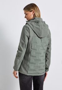 Lichtgrijs-groene jas met een capuchon, met een zachte buitenkant, een gewatteerd rugpaneel en zijzakken. De gestructureerde stof zorgt voor een gelaagd uiterlijk.