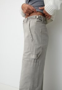 Pantaloni cargo grigio chiaro con più tasche, chiusure a bottone e cerniera, e una cintura robusta. Presentano una vestibilità comoda e un tessuto resistente.