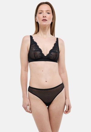 Femme portant un soutien-gorge en dentelle noire et une culotte transparente à pois, debout devant un fond blanc uni, face à l'avant.