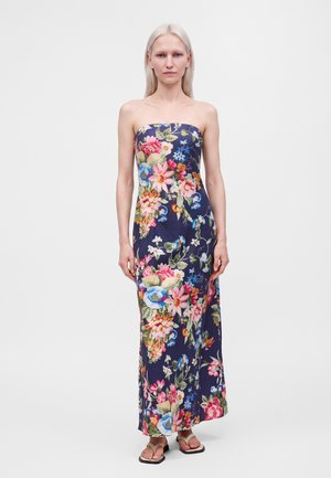 Donna con lunghi capelli biondi che indossa un vestito lungo blu navy senza spalline con stampa floreale colorata e sandali beige, in piedi su uno sfondo semplice.
