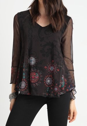Schwarze transparente Bluse mit langen Ärmeln, Blumenmuster in Rot, Blaugrün und Weiß am Saum, getragen mit schwarzer Hose und silbernen Armbändern.
