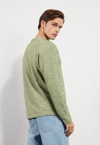 Chemise verte à manches longues avec une texture chinée, un col rond et une coupe décontractée, associée à un jean bleu clair.