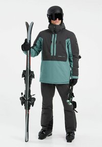 Skidragt bestående af en teal- og sort jakke med en lynlås lomme, kombineret med sorte bukser. Figuren holder ski og stave.