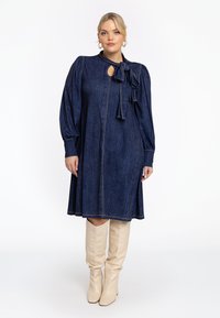 Dunkelblaudene A-Linien-Kleid mit Puffärmel, einem Schlüsselloch-Ausschnitt und Rüschendetail, ergänzt durch beigefarbene kniehohe Stiefel. Sichtbare Kontrastnähte.