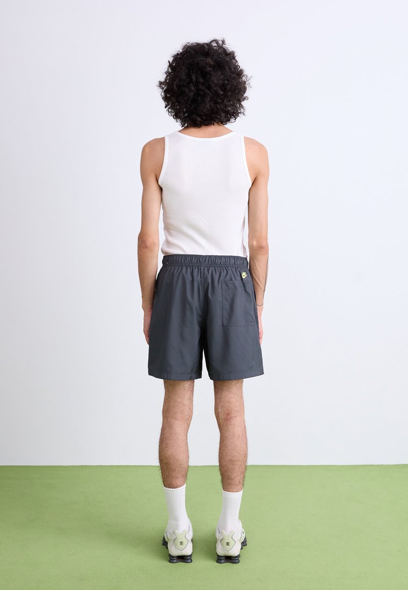 Pantaloni sportivi grigi con elastico in vita e tasca laterale, abbinati a una maglietta senza maniche bianca e calze bianche alla caviglia. Lo sfondo è chiaro.