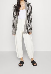 Cardigan à ouverture frontale gris avec motif en chevrons, associé à un pantalon large crème et des sandales slides noires. Matière en tricot texturé.