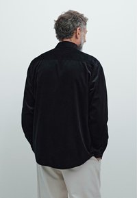 Sort fløjlsskjorte med afslappet pasform, der har en spredt krave og lange ærmer, set fra ryggen mod en lys grå baggrund.