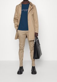 Man i beige kappa och byxor, marinblå T-shirt från Tommy Hilfiger, svarta stövlar, håller en svart läderhandväska framför en vit bakgrund.