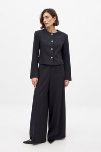 Melnā piegriezuma žakete ar zelta pogām, apvienota ar platiem melniem biksēm, kurām ir strukturēts dizains un gluda auduma tekstūra.