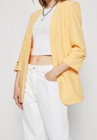 Gul kavaj med öppen framsidan och uppvuxna ärmar, parat med en vit crop top och hög midja vita jeans. Slät tygtextur.