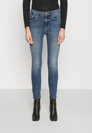 Smaltsiddende blå jeans med let falmning, båret med sorte ankelstøvler med hæl og en tætsiddende sort langærmet top, stående mod en ensfarvet baggrund.