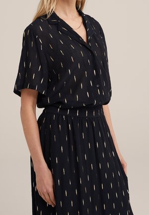 Vrouw die een zwarte blouse met korte mouwen en knopen draagt, en een bijpassende rok met een verticale gouden metalen stippenpatroon.