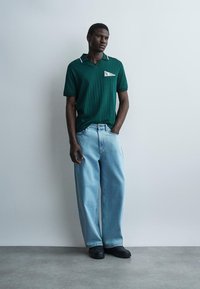 Άνδρας που φοράει σκούρο πράσινο κοντομάνικο πόλο και ανοιχτό μπλε τζιν με φαρδιά πόδια, στέκεται με το ένα χέρι στην τσέπη του απέναντι από έναν γκρίζο τοίχο.