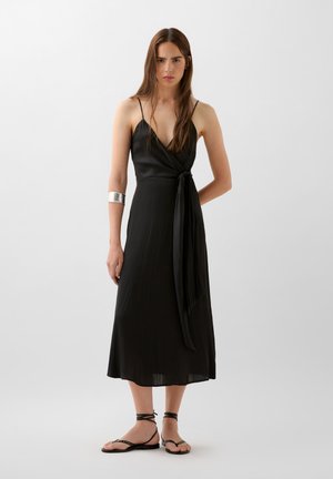 Black wrap dress with spaghetti straps, mid-length, made of smooth fabric. Features a tie at the waist and paired with flat sandals.