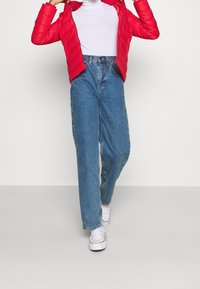 Rode gewatteerde pufferjack, hoge nek witte top, high-waisted lichtblauwe jeans en witte sneakers. Glad materiaal met een losse pasvorm.