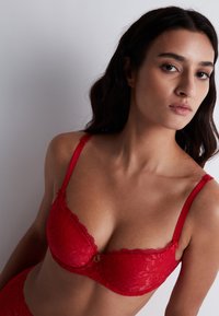 Soutien-gorge en dentelle rouge avec armatures, motif floral complexe, bords festonnés et bretelles ajustables. Présente un petit détail décoratif doré au centre.