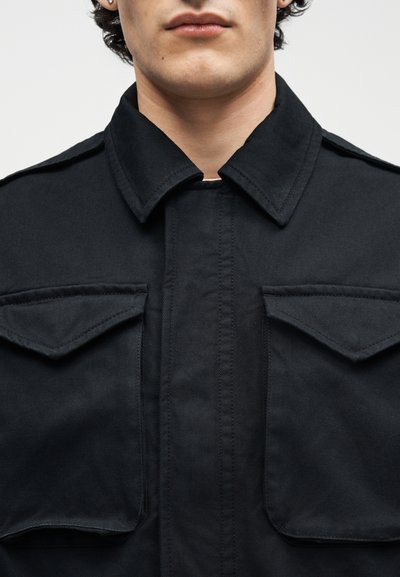 Κοντινό πλάνο ενός ατόμου που φορά μαύρο μπουφάν με φαρδιά γιακά και τσέπες με πτερύγια στο στήθος, εμφανίζοντας τον λαιμό και το κάτω μέρος του προσώπου.