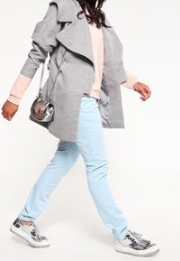 Cappotto lungo grigio con collo drappeggiato, maglione rosa chiaro sotto, pantaloni azzurri e scarpe da ginnastica argento con dettagli a frangia. Borsa argento.