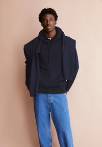 Marineblauer Pullover mit Kapuze und Vorderfach, getragen über einem schwarzen Hemd, kombiniert mit hellblauen Jeans aus Denim. Schlichtes Design, weiche Textur.