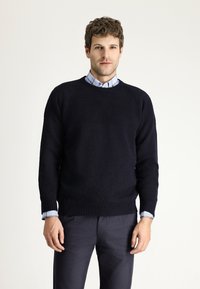 Marine gebreide trui met geribde mouwen en zoom, gedragen over een lichtblauw geblokt shirt. Soepele textuur en casual ontwerp.