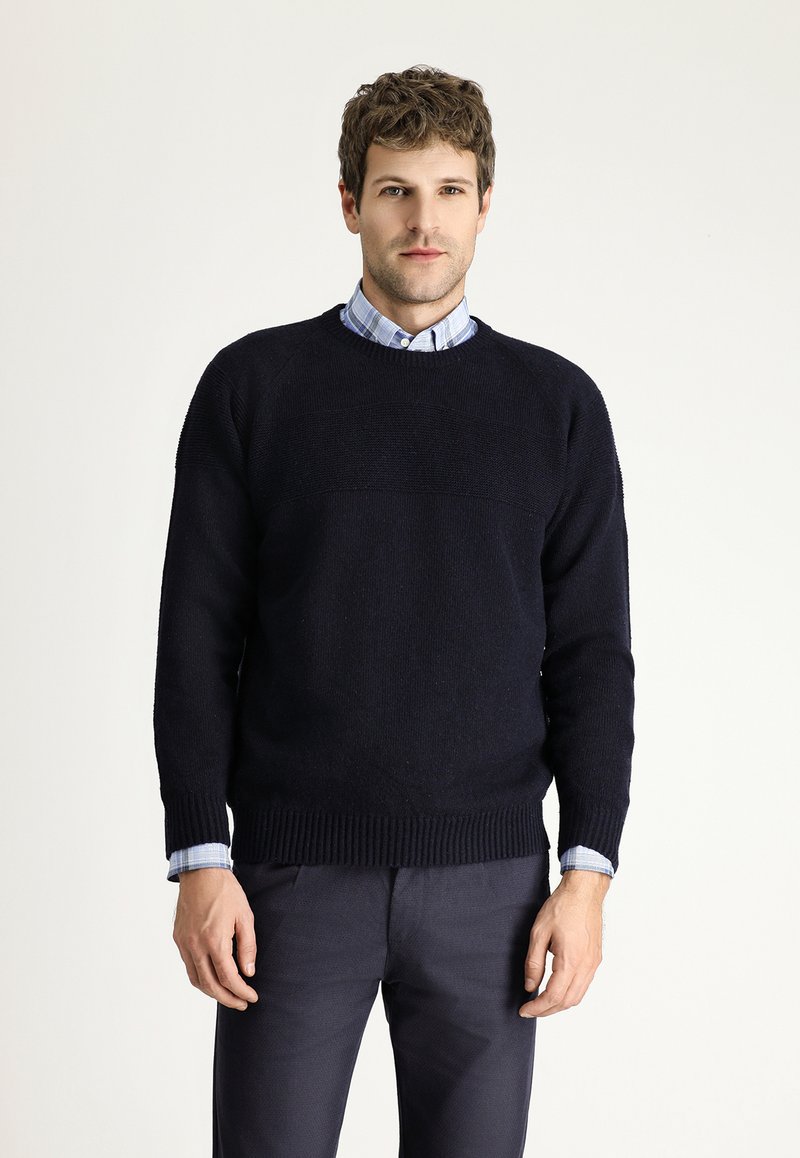 Marine gebreide trui met geribde mouwen en zoom, gedragen over een lichtblauw geblokt shirt. Soepele textuur en casual ontwerp.